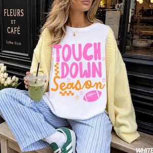 May include: White t-shirt with the text "TOUCH DOWN Season" in pink and orange, featuring a football graphic. The person is wearing a yellow cardigan, blue and white striped pants, and green sneakers. A clear cup with a green beverage is held in the hand.