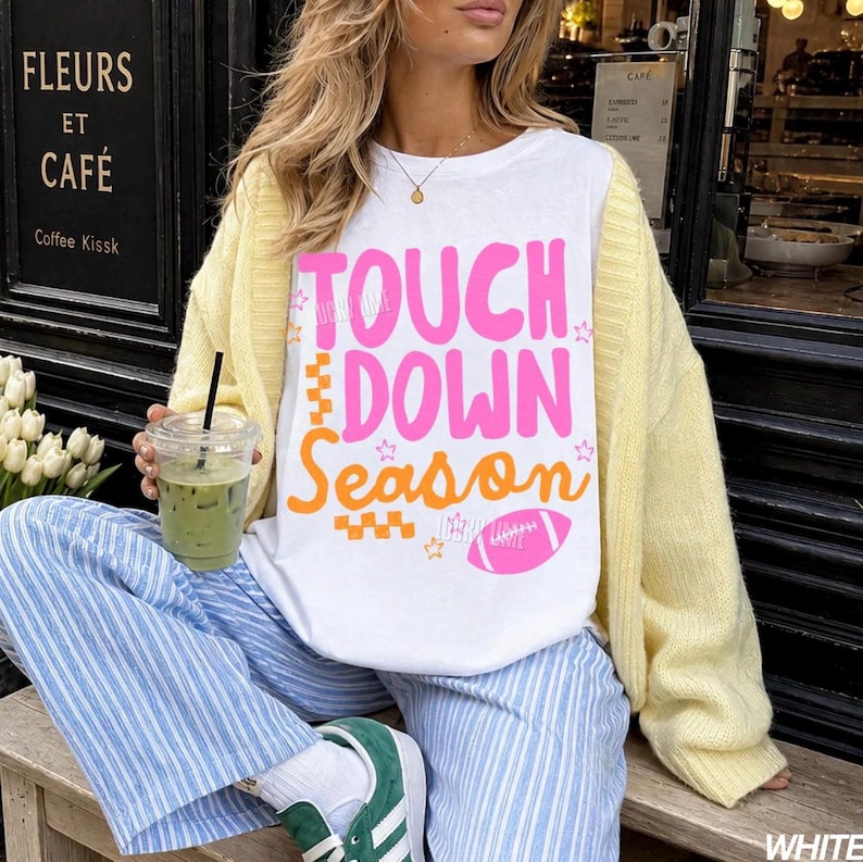 May include: White t-shirt with the text "TOUCH DOWN Season" in pink and orange, featuring a football graphic. The person is wearing a yellow cardigan, blue and white striped pants, and green sneakers. A clear cup with a green beverage is held in the hand.
