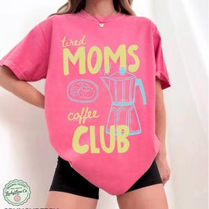 May include: A pink t-shirt with the words "tired MOMS coffee CLUB" in yellow and a light blue coffee pot graphic. The shirt also features a graphic of a croissant and a coffee pot. The shirt is a comfortable fit.