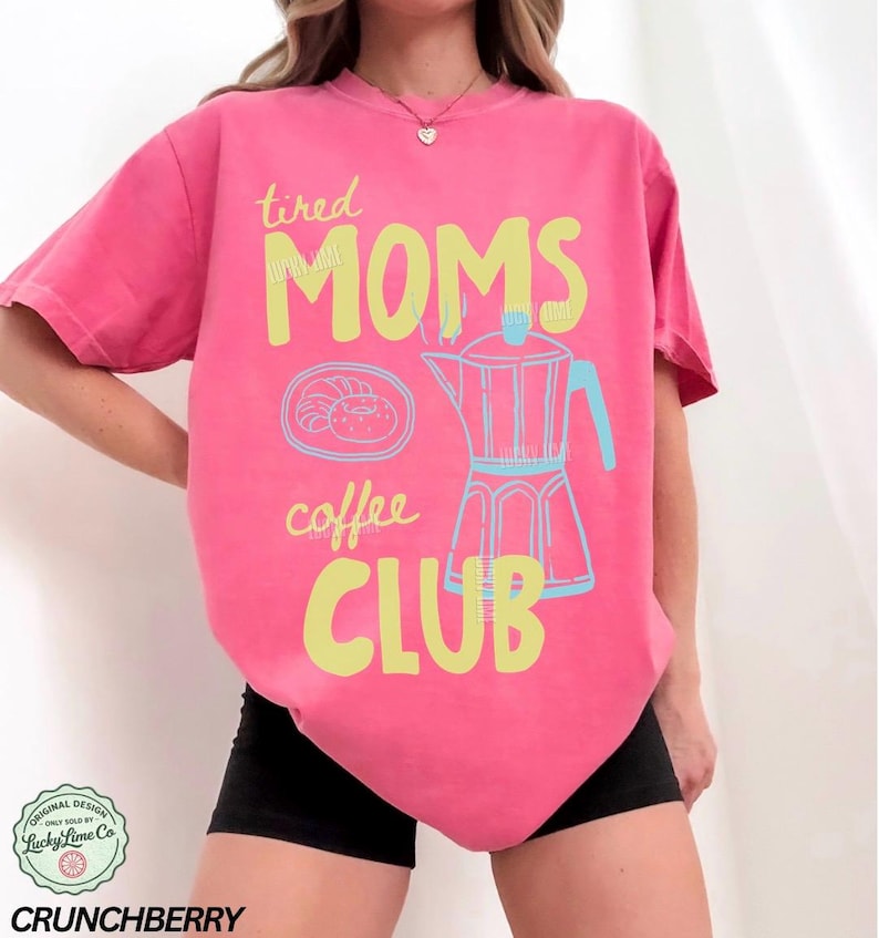 May include: A pink t-shirt with the words "tired MOMS coffee CLUB" in yellow and a light blue coffee pot graphic. The shirt also features a graphic of a croissant and a coffee pot. The shirt is a comfortable fit.