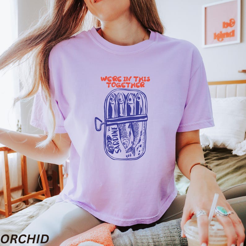 以下が含まれることがあります： 缶詰のサバと「We're in this together」というテキストが描かれたパープルのTシャツです。
