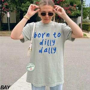 May include: A light green t-shirt with the text "born to dilly dally" in blue script. The shirt has a crew neck and short sleeves. The shirt is a relaxed fit and appears to be made of a soft material.