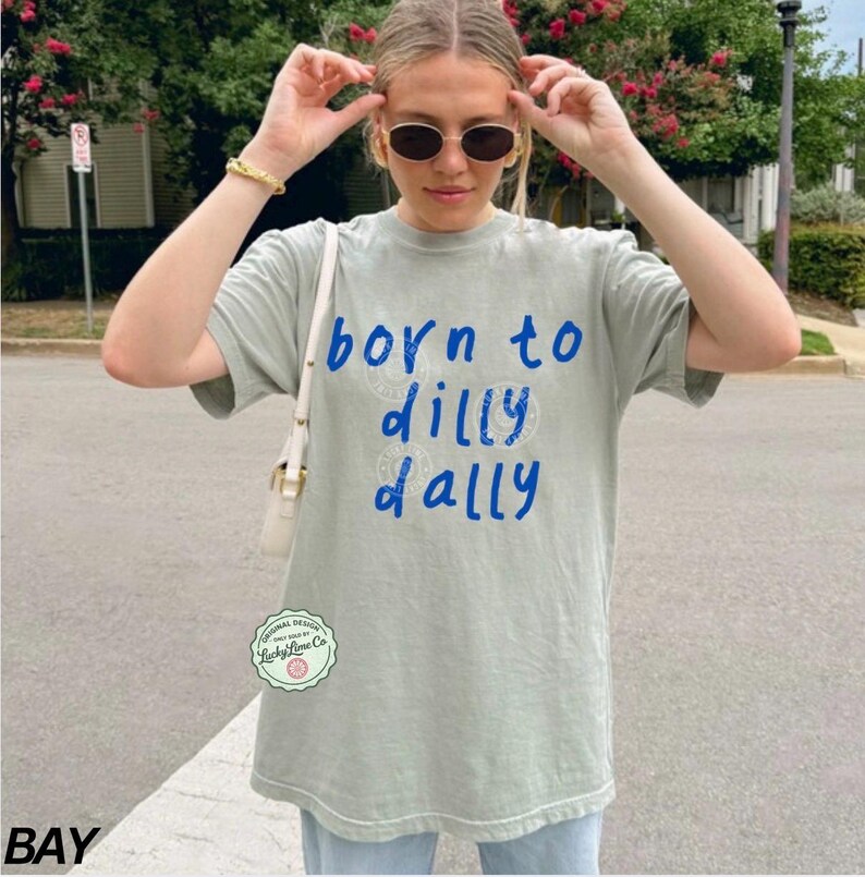 May include: A light green t-shirt with the text "born to dilly dally" in blue script. The shirt has a crew neck and short sleeves. The shirt is a relaxed fit and appears to be made of a soft material.