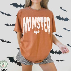May include: Burnt orange t-shirt with the word "MOMSTER" in white, featuring a bat graphic. The shirt is part of a Halloween-themed collection. The model is wearing black and white checkered shorts.