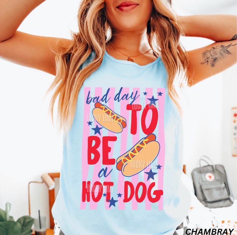 Op de afbeelding: Lichtblauw tanktop met de tekst "bad day to be a hot dog" in rood en blauw. Het ontwerp bevat twee hotdogs en sterren, met roze en witte strepen op de achtergrond.