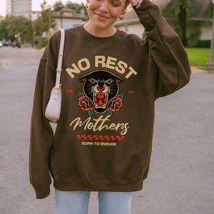 Op de afbeelding: Een bruin sweatshirt met een zwarte panter afbeelding en de tekst "No Rest for the Mothers Born to Endure".