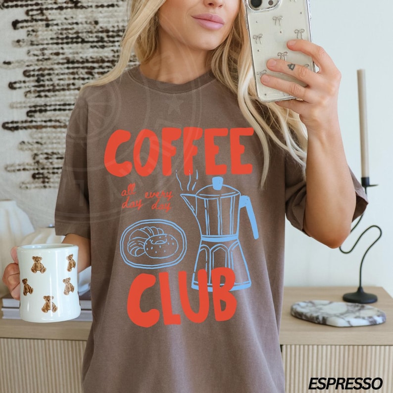 Op de afbeelding: Een bruin T-shirt met de tekst "COFFEE CLUB" in rood en blauw. De tekst "all day every day" staat in kleinere letters onder het woord "COFFEE". Een blauwe en witte illustratie van een koffiekan en gebakjes bevindt zich in het midden van het shirt.