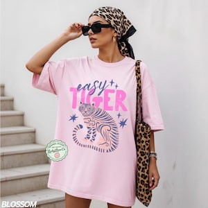 Puede incluir: Camiseta rosa claro con la frase "easy TIGER" en rosa y una ilustraci&oacute;n de tigre. La camiseta es parte de la colecci&oacute;n "BLOSSOM". Tambi&eacute;n se ve un bolso tote y una bufanda con estampado de leopardo.