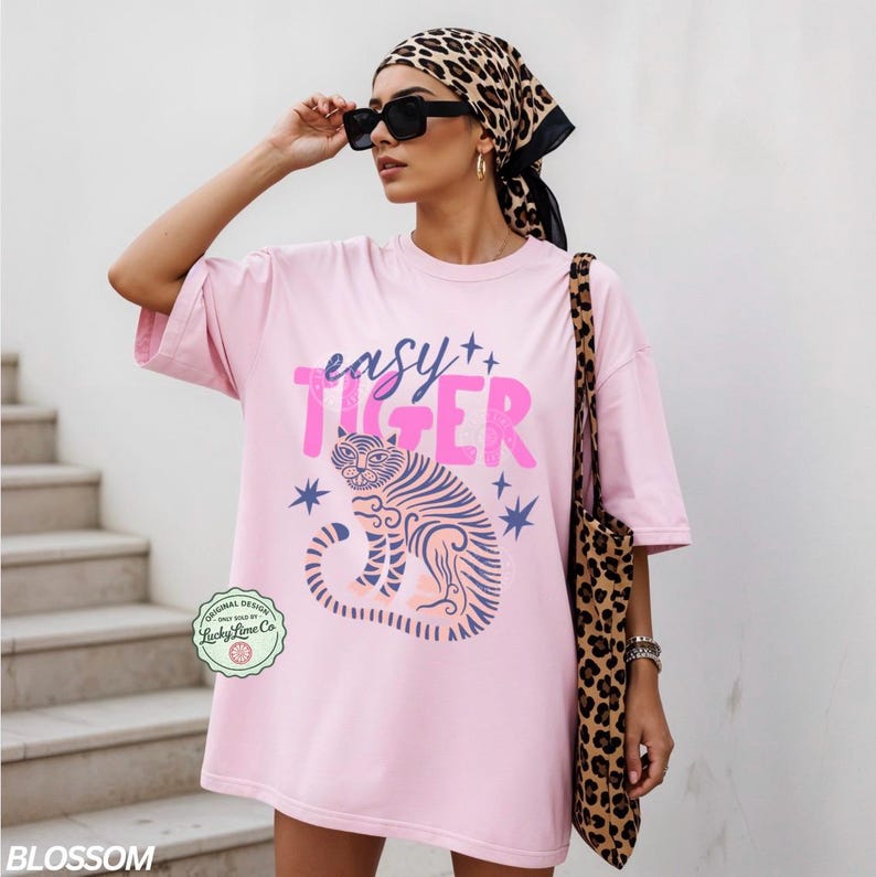 Puede incluir: Camiseta rosa claro con la frase "easy TIGER" en rosa y una ilustraci&oacute;n de tigre. La camiseta es parte de la colecci&oacute;n "BLOSSOM". Tambi&eacute;n se ve un bolso tote y una bufanda con estampado de leopardo.