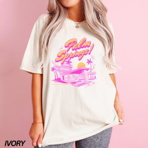May include: A white t-shirt featuring a pink and orange graphic design that reads "Palm Springs!" and depicts a retro-style motel building with palm trees and a sunset. The shirt is available in ivory.