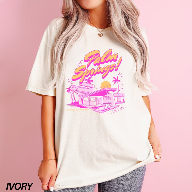 May include: A white t-shirt featuring a pink and orange graphic design that reads "Palm Springs!" and depicts a retro-style motel building with palm trees and a sunset. The shirt is available in ivory.