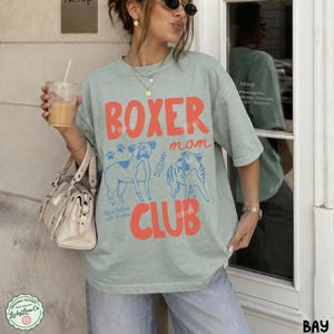 May include: A sage green short-sleeve t-shirt featuring the red text BOXER MOM CLUB and blue line art illustrations of two boxer dogs. The design includes small paw prints and the text Life is better with boxers in a script font.