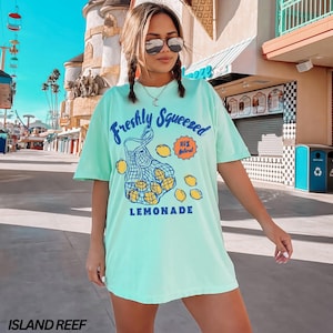 Puede incluir: Una camiseta azul turquesa con un gr&aacute;fico de una bolsa de malla llena de limones y el texto "Freshly Squeezed 100% Natural LEMONADE".