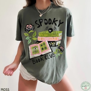 Op de afbeelding: Mosgroen t-shirt met een "Spooky Book Club" ontwerp. De afbeelding toont een spin op boeken, open boeken met illustraties en zwarte rozen. Het shirt heeft een relaxte pasvorm.