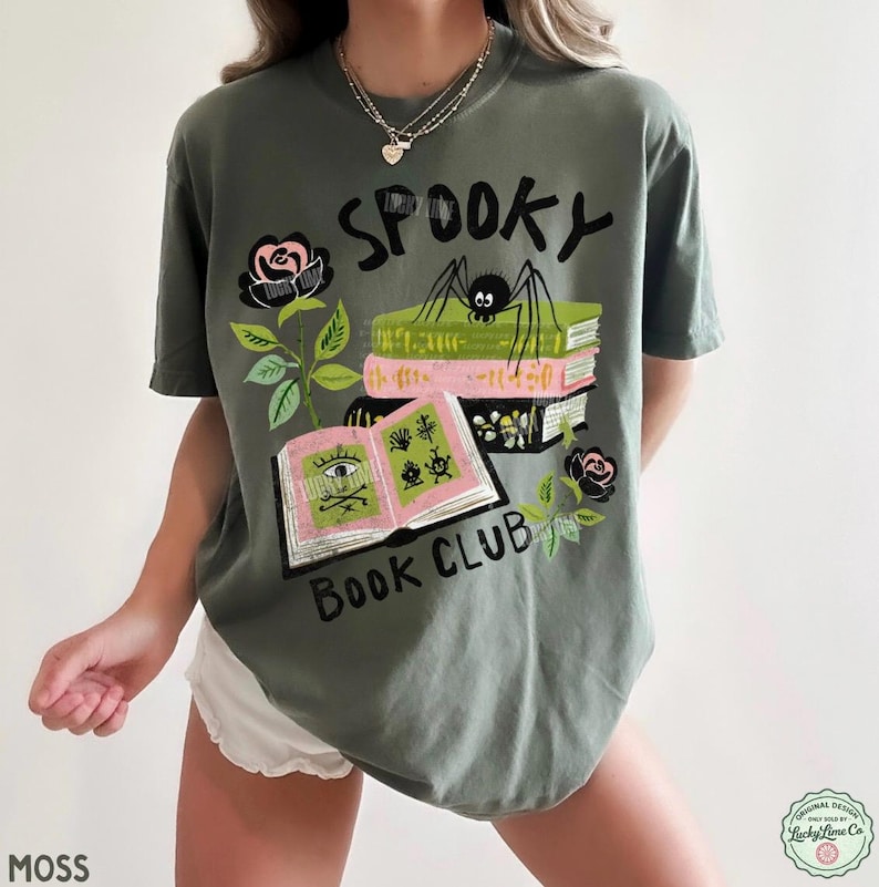 Op de afbeelding: Mosgroen t-shirt met een "Spooky Book Club" ontwerp. De afbeelding toont een spin op boeken, open boeken met illustraties en zwarte rozen. Het shirt heeft een relaxte pasvorm.