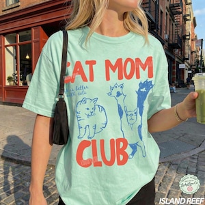 Puede incluir: Una camiseta verde claro con las palabras "CAT MOM CLUB" en rojo. La camiseta presenta ilustraciones de gatos azules y la frase "is better with cats". La camiseta es de estilo casual y c&oacute;modo.