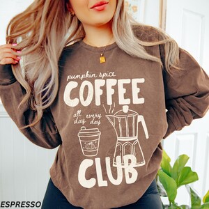 May include: A brown sweatshirt with the text "pumpkin spice COFFEE CLUB all every day day". The design includes a coffee cup with a pumpkin and a coffee maker. The sweatshirt is a warm, casual style.