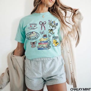 May include: A pale turquoise t-shirt with a graphic design featuring a teacup, cake, books, butterflies, flowers, and a jar with lemons. The text "CHALKY MINT" is printed in the bottom right corner.