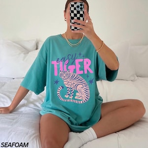 Puede incluir: Camiseta verde mar con la frase "easy tiger" en rosa sobre una ilustraci&oacute;n de tigre rosa y morado. La camiseta tiene mangas cortas y un corte holgado.