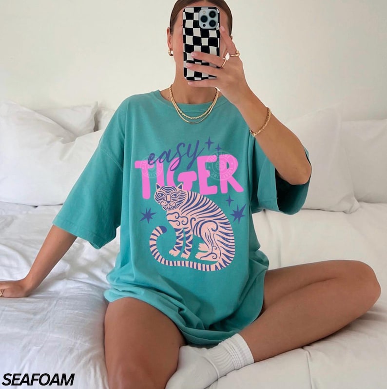 Puede incluir: Camiseta verde mar con la frase "easy tiger" en rosa sobre una ilustraci&oacute;n de tigre rosa y morado. La camiseta tiene mangas cortas y un corte holgado.