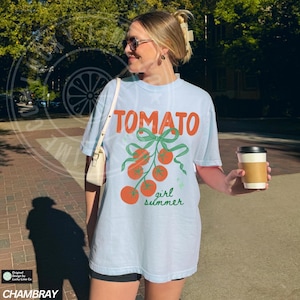 May include: Light blue t-shirt with the word "TOMATO" in orange, and a graphic of tomatoes with green bows and the words "girl summer" in green. The shirt is made of a soft material.