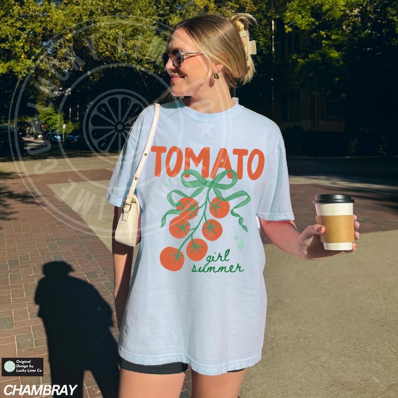 May include: Light blue t-shirt with the word "TOMATO" in orange, and a graphic of tomatoes with green bows and the words "girl summer" in green. The shirt is made of a soft material.