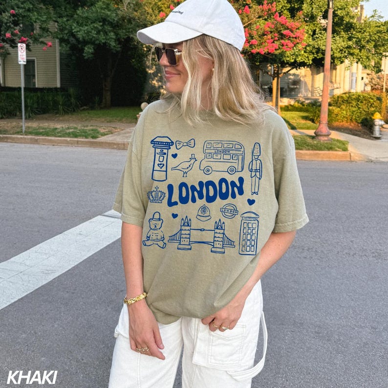 May include: A khaki t-shirt with a blue graphic print of London landmarks, including a double-decker bus, a phone booth, and Big Ben. The text "LONDON" is printed in a large, bold font.
