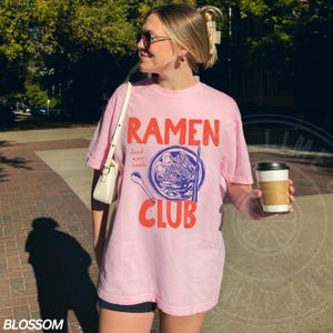 May include: Pink t-shirt with a graphic of a bowl of ramen noodles and the text "Ramen Club" in red and blue. The text "Send more noodles" is written in smaller blue text.