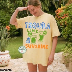 May include: A pale yellow t-shirt with the text "TEQUILA LIME & SUNSHINE" in blue, green, and orange. The shirt displays a graphic of two cocktails with lime slices. The text "ORIGINAL DESIGN" and "Lucky Lime Co" are also visible.