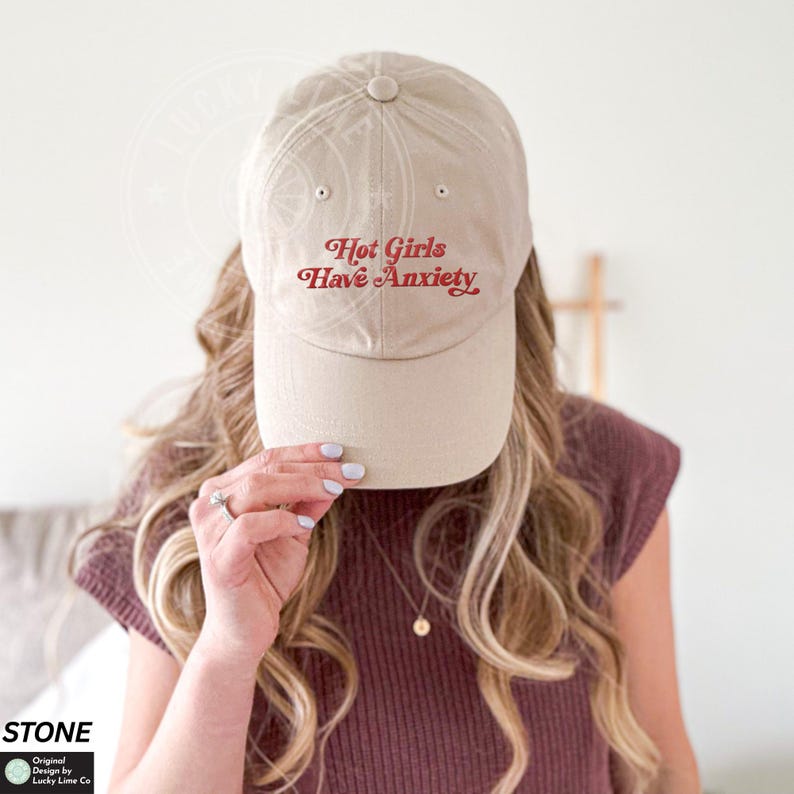 May include: A beige baseball cap with the text "Hot Girls Have Anxiety" in red script. The cap is made of a soft material and has a curved brim. The hat is a casual accessory.