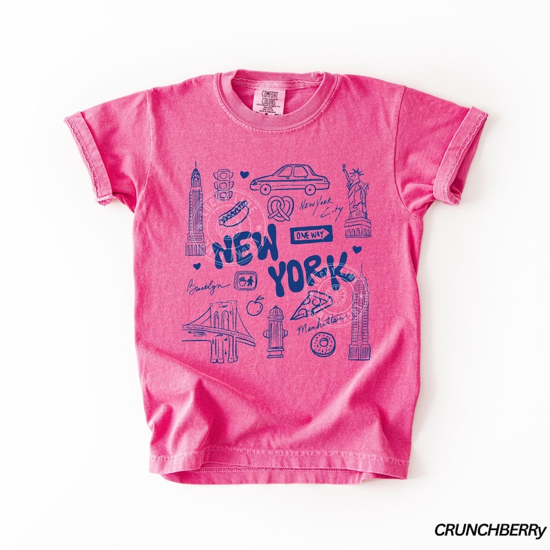 May include: A pink t-shirt with a blue graphic design featuring New York City landmarks and the text "NEW YORK". The design includes illustrations of the Statue of Liberty, the Empire State Building, and a pretzel.