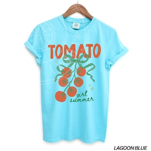 May include: A lagoon blue t-shirt with rolled-up sleeves. The shirt features the word "TOMATO" in orange block letters above a green bow and a cluster of orange tomatoes. Below the tomatoes, the words "girl summer" are written in green script.