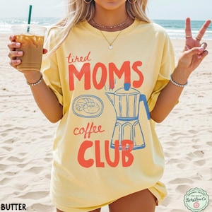 May include: A pale yellow t-shirt featuring the red text "tired MOMS" and "coffee CLUB" alongside a blue graphic of a moka pot and a plate with a pastry. The design is centered on the front of the shirt, which is worn in a beach setting.