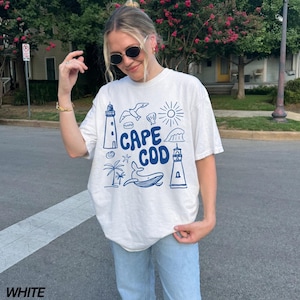 Peut inclure: T-shirt blanc avec un motif graphique bleu qui dit "Cape Cod" et repr&eacute;sente un phare, une baleine, une mouette et d'autres &eacute;l&eacute;ments nautiques.