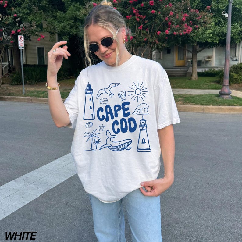 Peut inclure: T-shirt blanc avec un motif graphique bleu qui dit "Cape Cod" et repr&eacute;sente un phare, une baleine, une mouette et d'autres &eacute;l&eacute;ments nautiques.