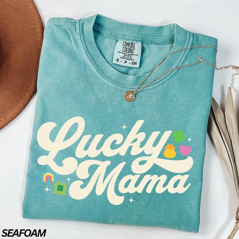 Peut inclure: Un t-shirt bleu turquoise avec le texte "Lucky Mama" dans une police r&eacute;tro. Le texte est de couleur cr&egrave;me et est entour&eacute; de petits graphiques de tr&egrave;fles et d'arcs-en-ciel.