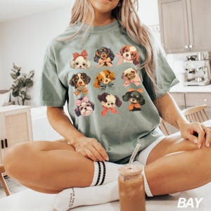 May include: A green t-shirt with a graphic print of 10 different dogs wearing bows. The dogs are all different breeds and have different expressions. The shirt is being worn by a person sitting on a chair.