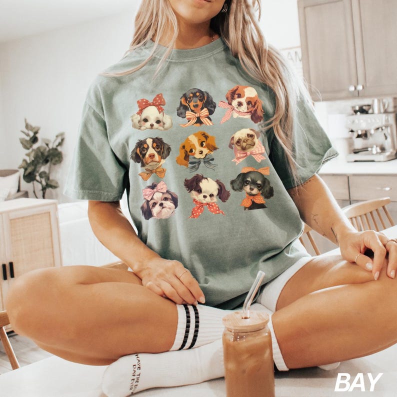 May include: A green t-shirt with a graphic print of 10 different dogs wearing bows. The dogs are all different breeds and have different expressions. The shirt is being worn by a person sitting on a chair.