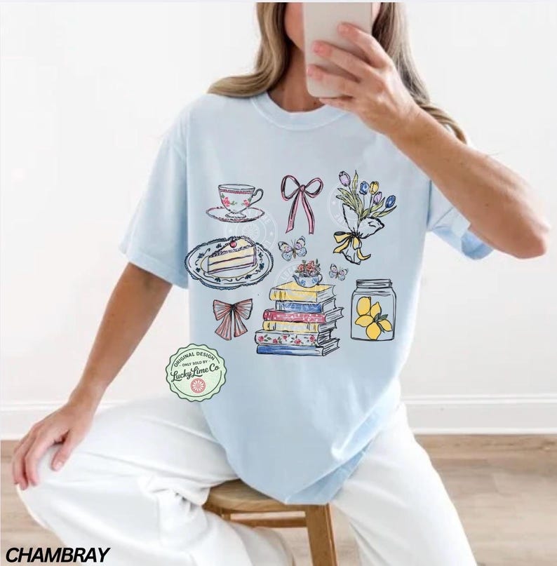 May include: Pale blue t-shirt with a charming illustration of a teacup, cake, bows, butterflies, flowers, books, and a lemon jar. The shirt has the text "Original Design" and "Lucky Lime Co."