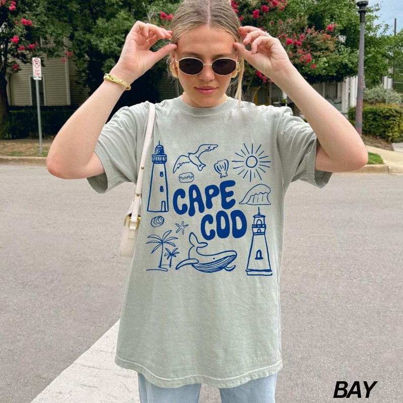 Peut inclure: Un t-shirt vert clair avec un motif graphique bleu repr&eacute;sentant un phare, une baleine, une mouette, une coquille, un palmier, un soleil et une vague. Le texte "CAPE COD" est &eacute;crit en bleu sur le t-shirt.