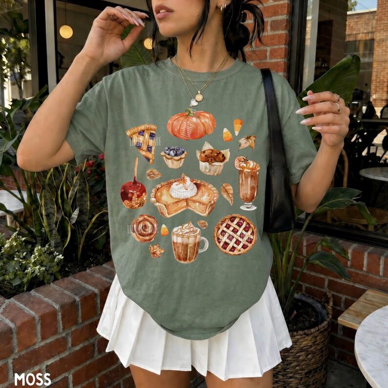 May include: An olive green t-shirt featuring a graphic print of various autumn-themed desserts and treats, including a pumpkin, a slice of pie, a caramel apple, a muffin, a latte, and a lattice-crust pie, paired with a white pleated mini skirt.