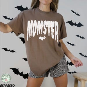 May include: A brown t-shirt with the word "MOMSTER" in white, with a bat graphic. The shirt is part of a Halloween-themed collection. The model is wearing black and white checkered shorts.