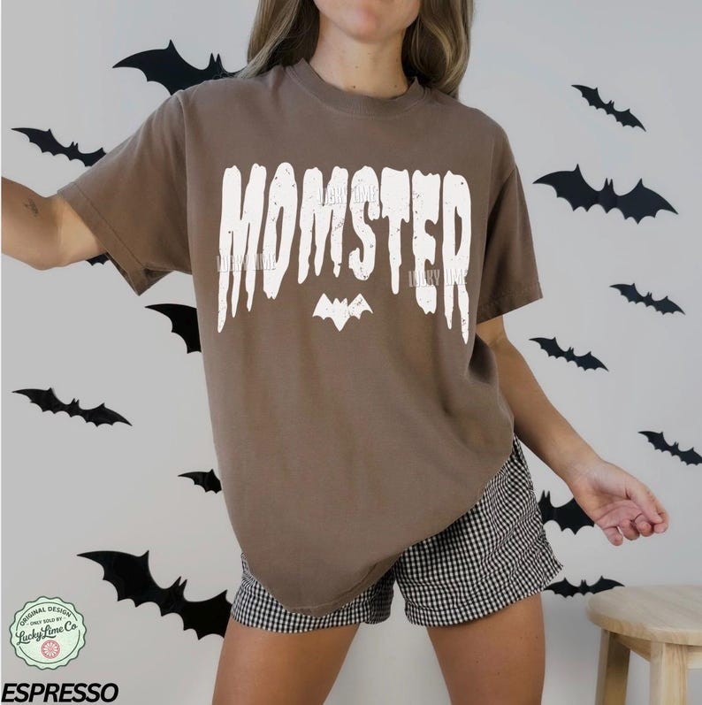 May include: A brown t-shirt with the word "MOMSTER" in white, with a bat graphic. The shirt is part of a Halloween-themed collection. The model is wearing black and white checkered shorts.