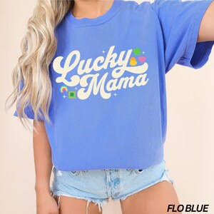 Peut inclure: Un t-shirt bleu avec le texte "Lucky Mama" dans une police r&eacute;tro. Le texte est en blanc avec une pi&egrave;ce d'or jaune, un tr&egrave;fle vert et des accents arc-en-ciel.
