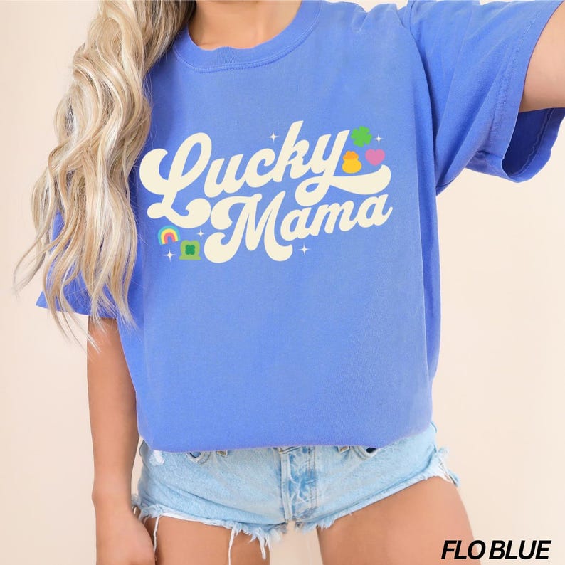 Peut inclure: Un t-shirt bleu avec le texte "Lucky Mama" dans une police r&eacute;tro. Le texte est en blanc avec une pi&egrave;ce d'or jaune, un tr&egrave;fle vert et des accents arc-en-ciel.
