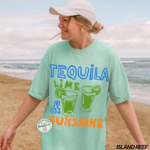 May include: A pale green t-shirt with the words "TEQUILA LIME & SUNSHINE" in blue and orange. The shirt features two green cocktail glasses with lime slices. The design includes a small circular logo that says "Lucky Lime Co."