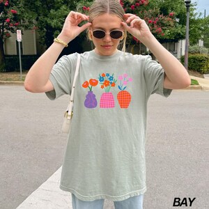 May include: A light green t-shirt with a graphic of colorful flowers in vases. The vases are purple, pink, and orange with geometric patterns.