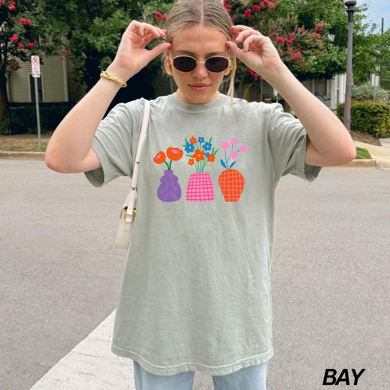 May include: A light green t-shirt with a graphic of colorful flowers in vases. The vases are purple, pink, and orange with geometric patterns.
