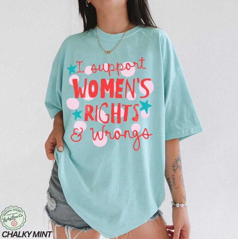 Peut inclure: T-shirt bleu clair avec l'inscription "I support WOMEN'S RIGHTS & Wrongs" en rouge et rose. Le t-shirt a une coupe ample et des manches courtes. Le texte est orn&eacute; de pois roses et d'&eacute;toiles bleues.
