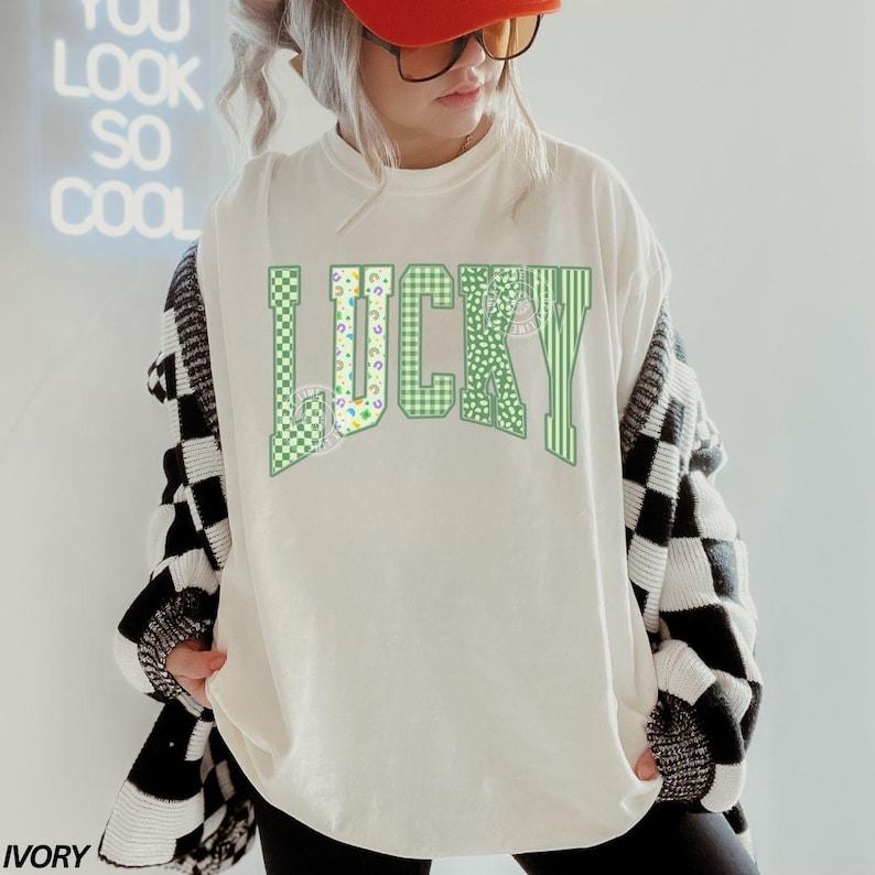 May include: Ivory-colored t-shirt with the word "LUCKY" in green, featuring various patterns. The shirt is paired with a black and white checkered cardigan. The word "IVORY" is printed at the bottom of the shirt.